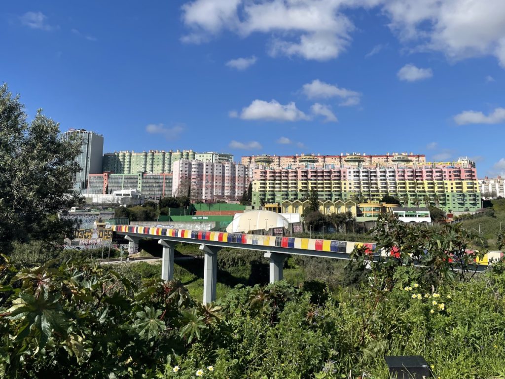 Kilátás Lisszabon egyik metróvonal-hídjára. A háttérben színes magas tömbházak. A táj tele van zöld növényzettel.