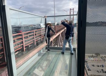 Turista kapaszkodik a lengőhíd üvegfalára. A párja fotózza.